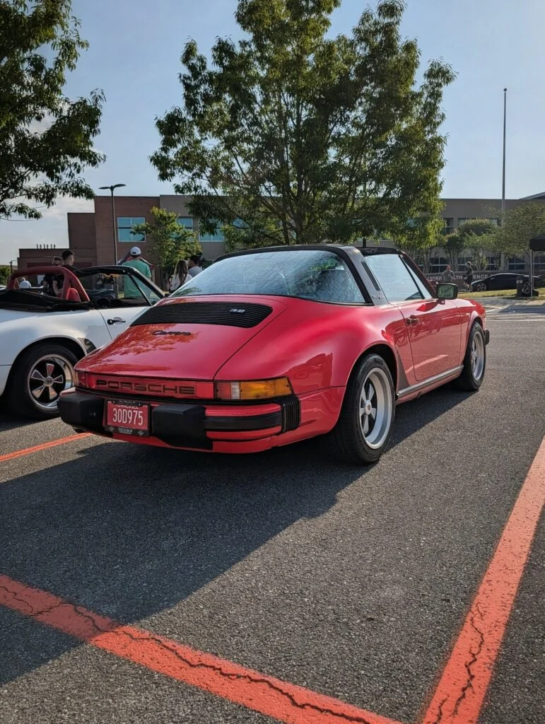 Red Porsche 911 Targa parked at an outdoor car event with people and a white convertible nearby under trees and clear sky.