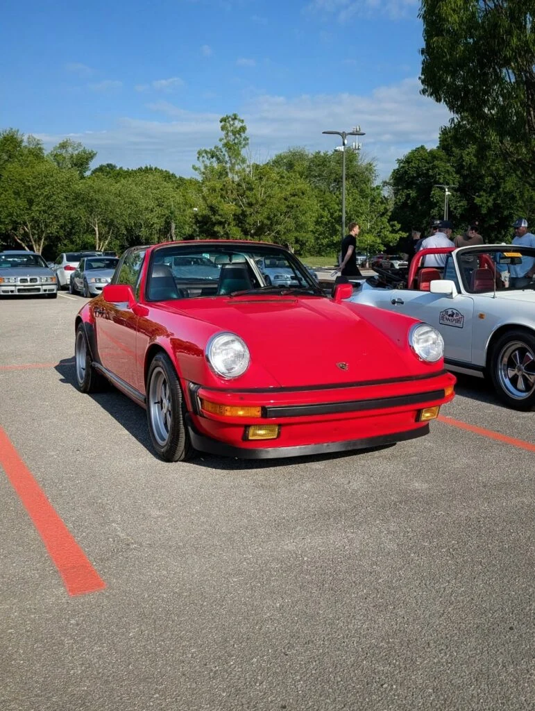 Red Porsche 911 Targa in a parking lot at a car event on a sunny day with other cars and people in background