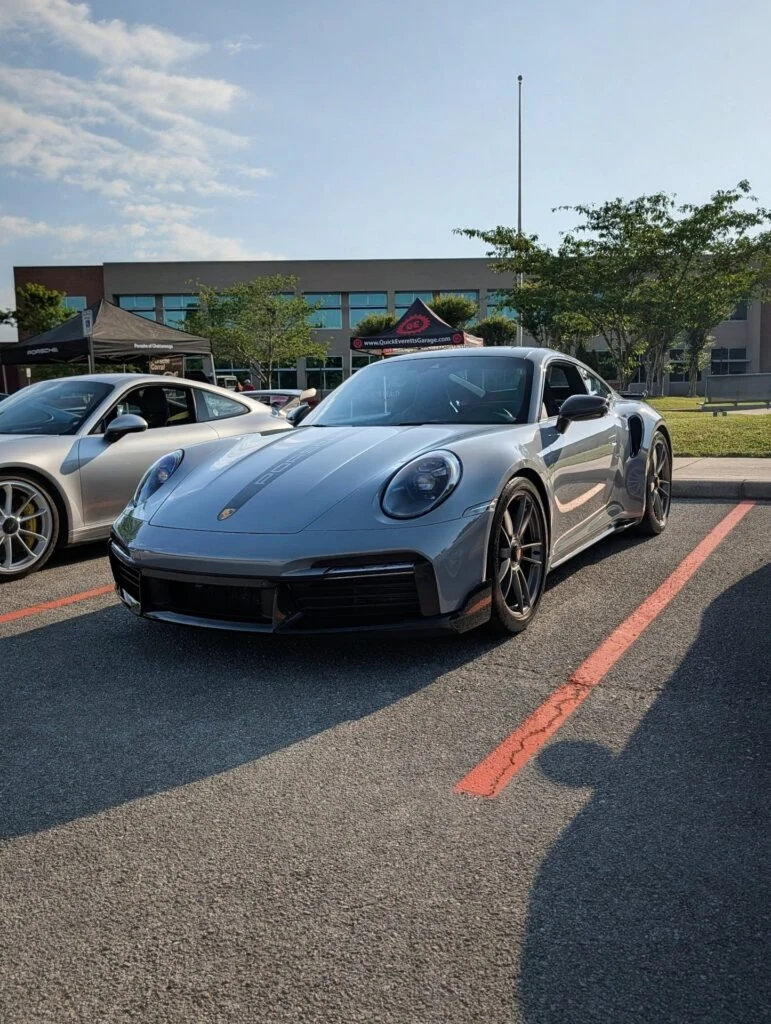 Silver Porsche 911 Turbo S parked at an outdoor car event on a sunny day with tents and trees in the background.