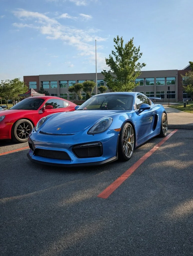 Blue Porsche 718 Cayman GT4 and red Toyota 86 parked outdoors in a parking lot near a building with trees nearby.