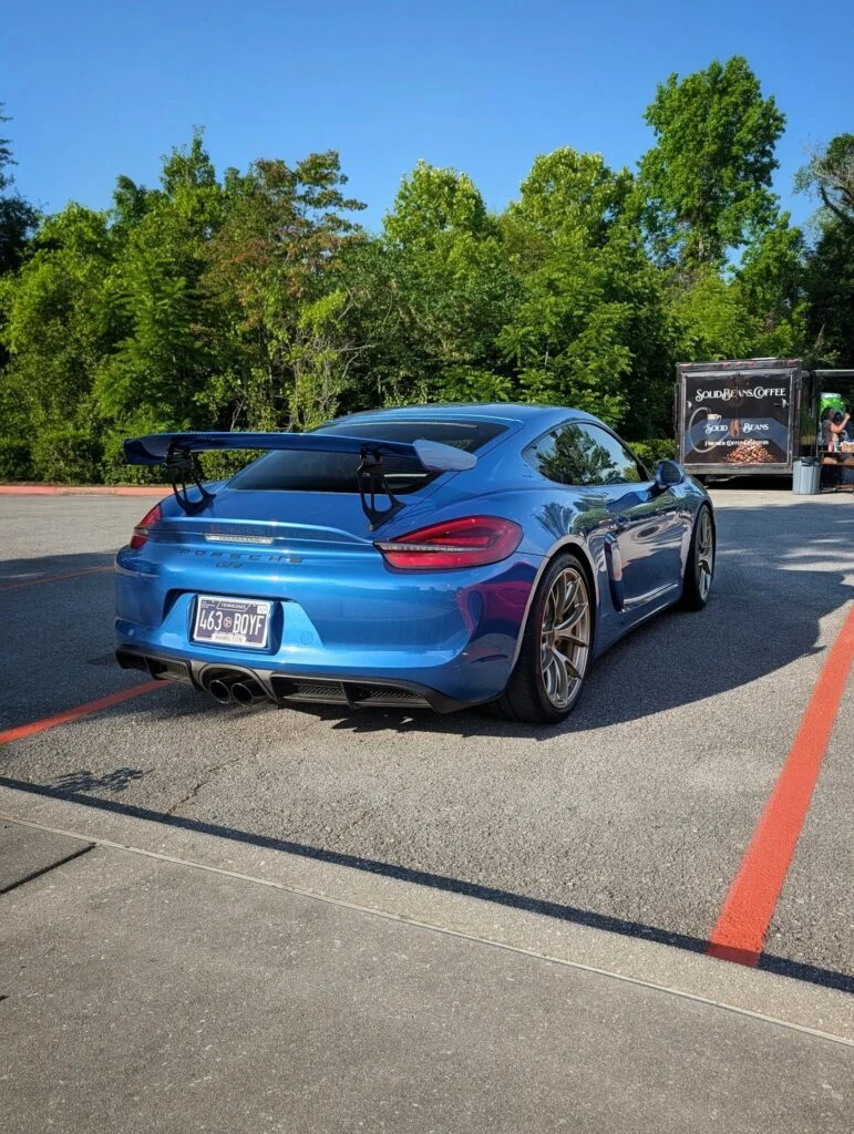 Blue Porsche 718 Cayman GT4 parked in a lot near green trees and a coffee vendor truck