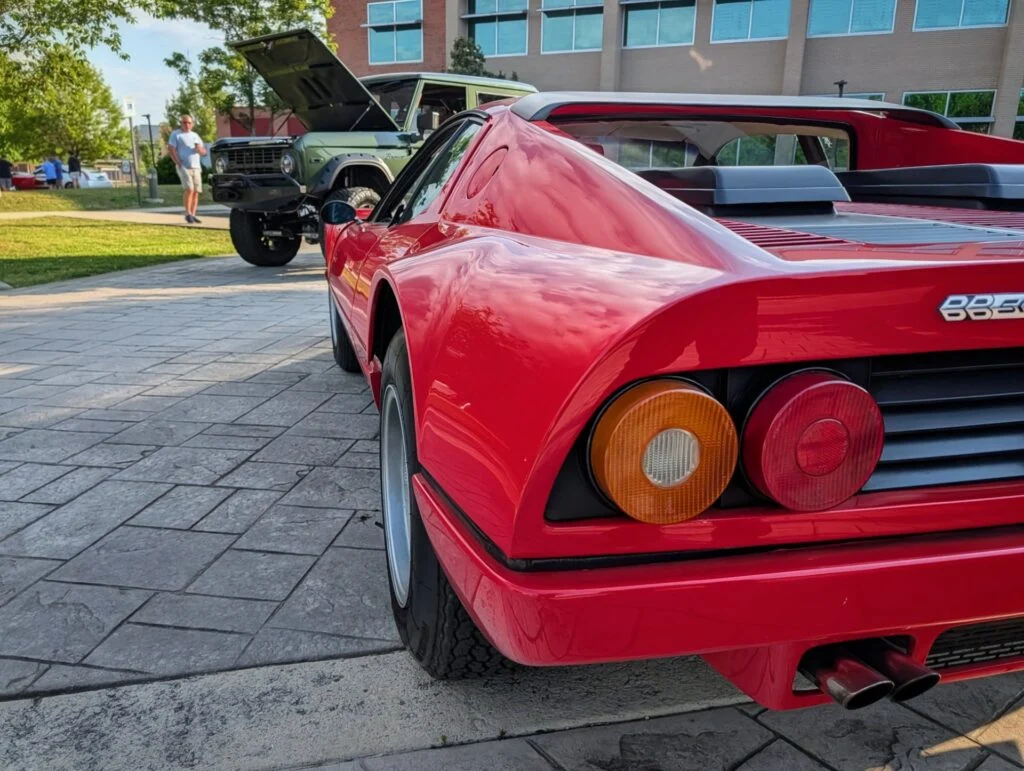 Red Ferrari BB 512 rear 3/4 view parked near an olive green Ford Bronco with its hood open at an outdoor car event.