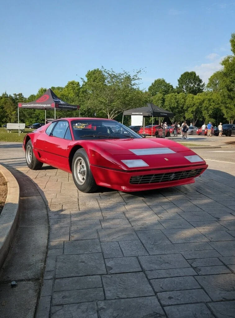 Red Ferrari 512 BB parked in an outdoor area with event tents and people in the background under clear blue sky