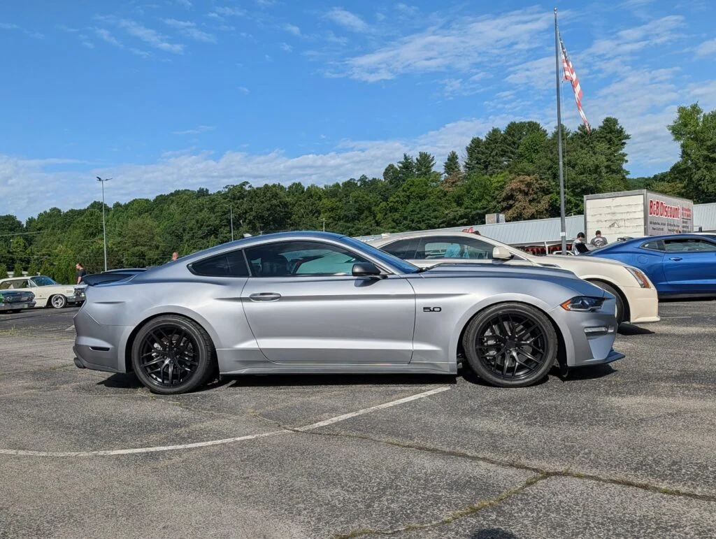 Silver Ford Mustang 5.0 parked in a lot with other cars and people visible in the background under a blue sky