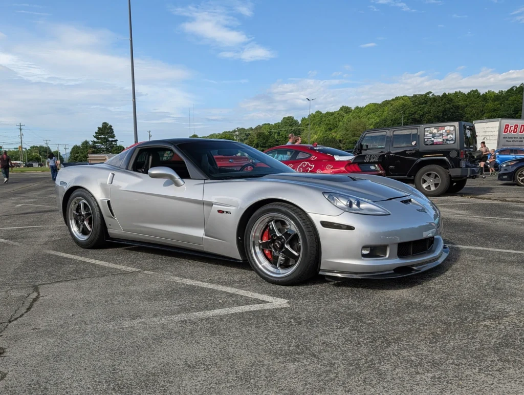 Silver Chevrolet Corvette Z06 parked in a lot with other vehicles and people walking nearby under a partly cloudy sky