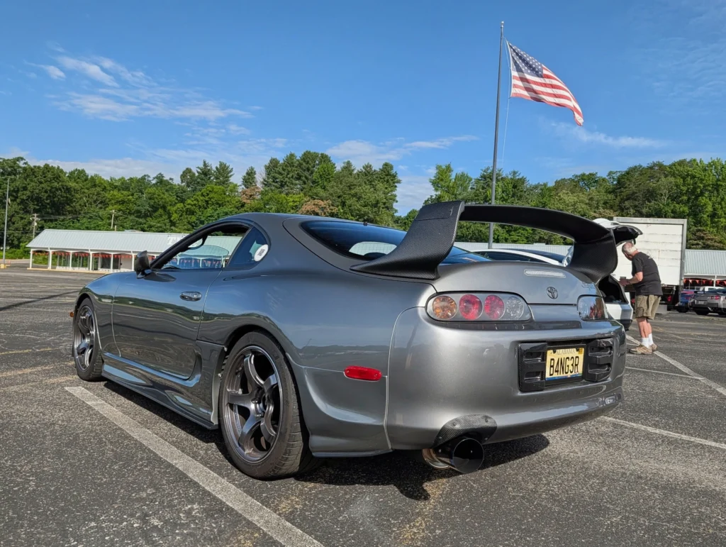 Silver Toyota Supra with large rear wing in a parking lot with an American flag in the background