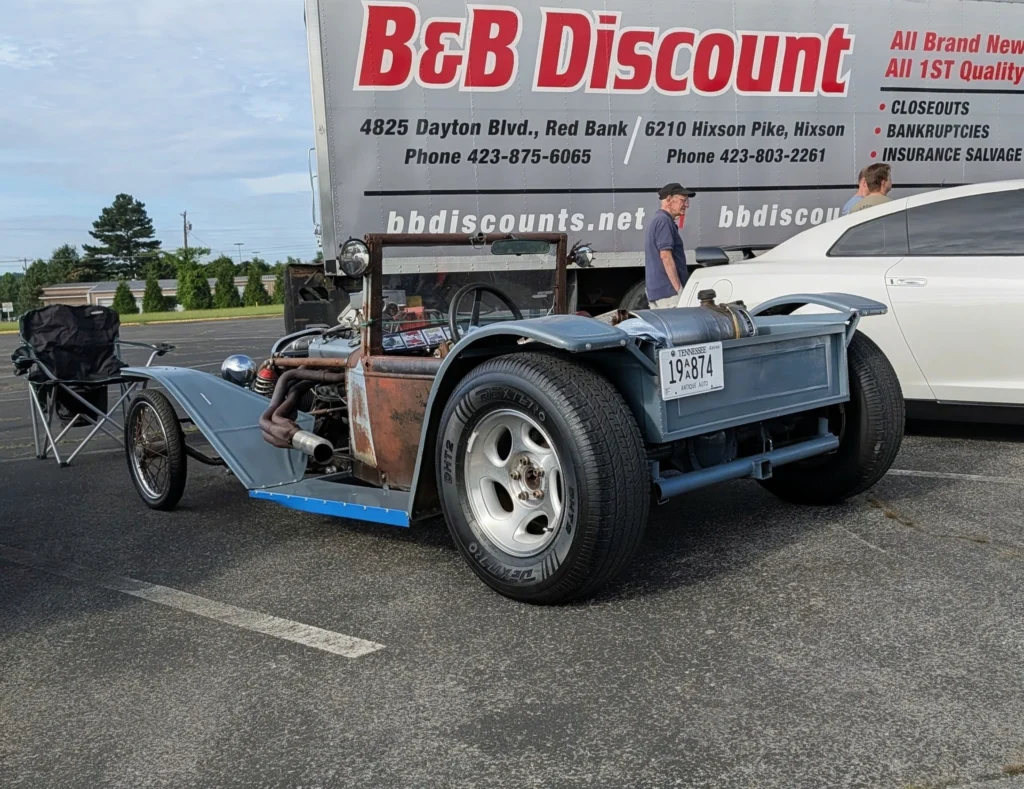 Rusty homemade hot rod with Tennessee antique license plate parked next to a white car and a man standing in a parking lot ne