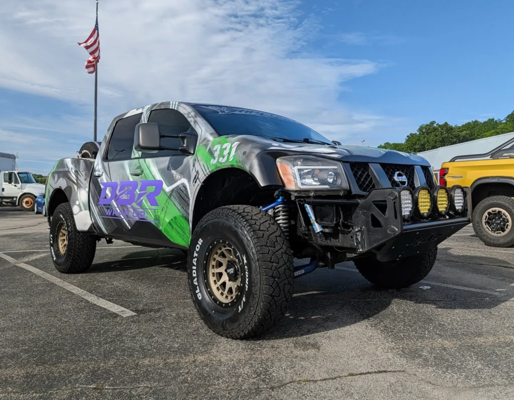 Gray and green Nissan off-road truck parked in a car event lot under a partly cloudy sky with an American flag in the backgro