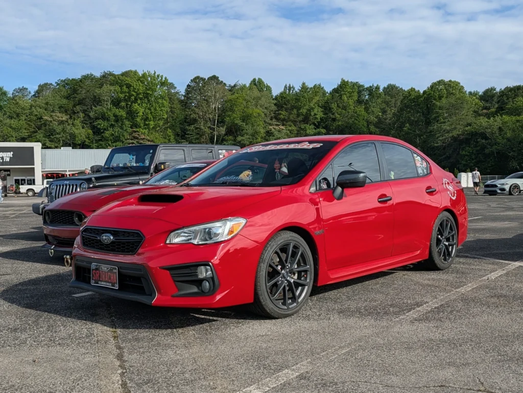Red Subaru WRX parked next to a brown Dodge Challenger and a black Jeep in a parking lot with trees and building in backgroun