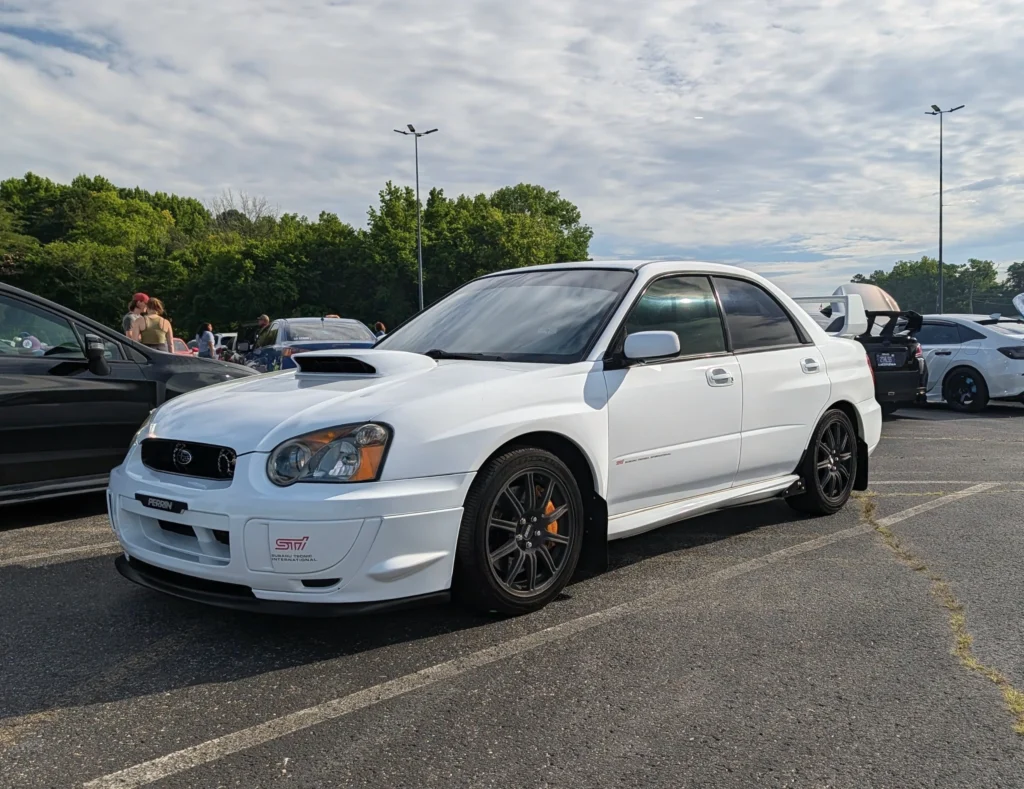 White Subaru Impreza WRX STI parked in a lot with other cars and people in the background under a partly cloudy sky