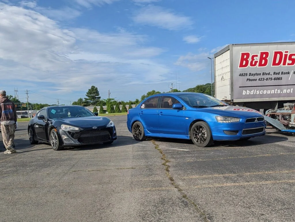 Black Scion coupe and blue Mitsubishi Lancer parked on asphalt near a truck trailer in a parking lot under a blue sky with clo