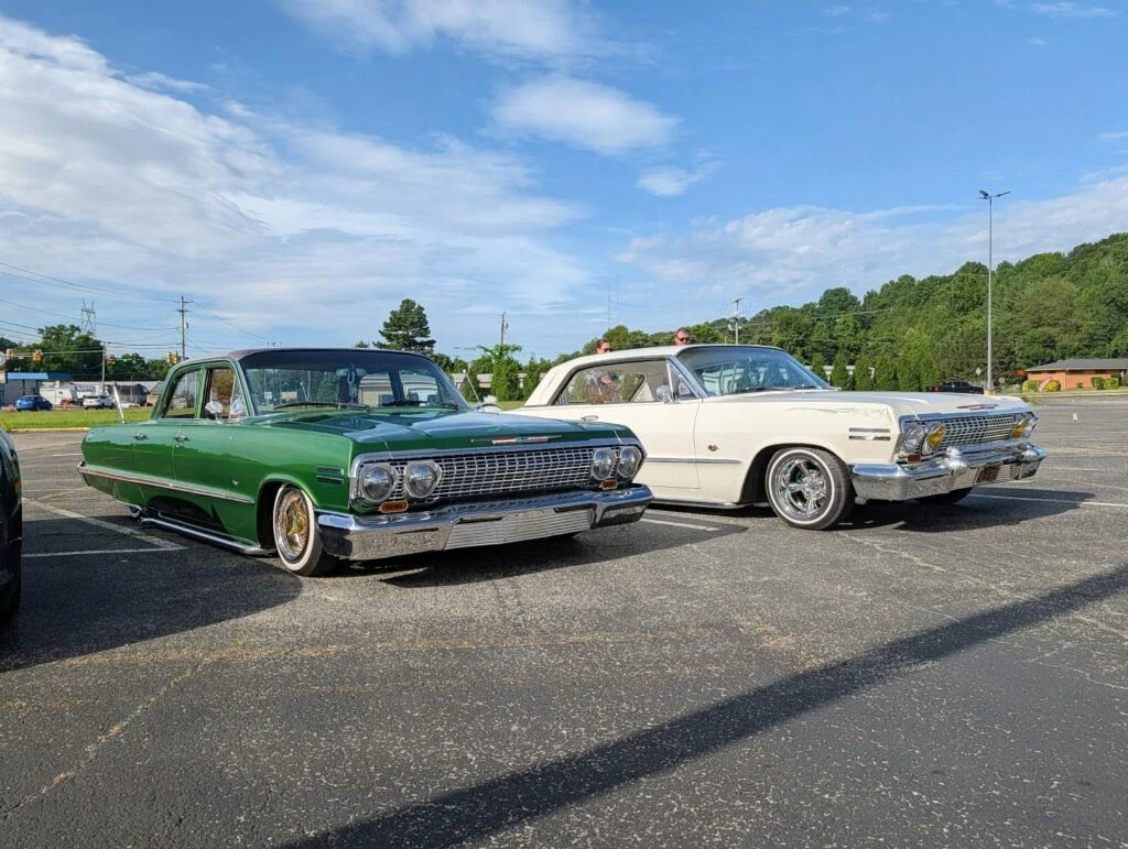 Green and white Chevrolet classic cars parked side by side in an outdoor parking lot under a partly cloudy sky