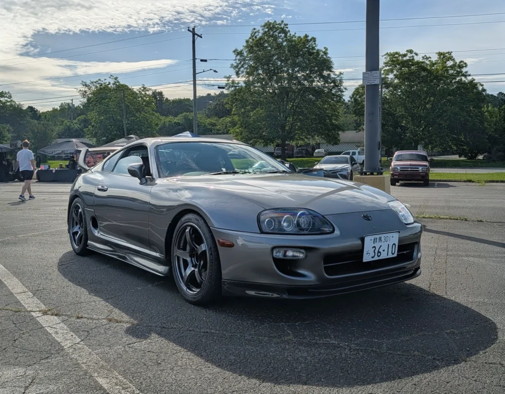 Gray Toyota Supra parked in a lot with tents and people in the background under a partly cloudy sky