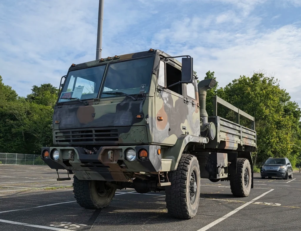 Large camouflaged military truck parked in a lot with trees and a silver SUV in the background