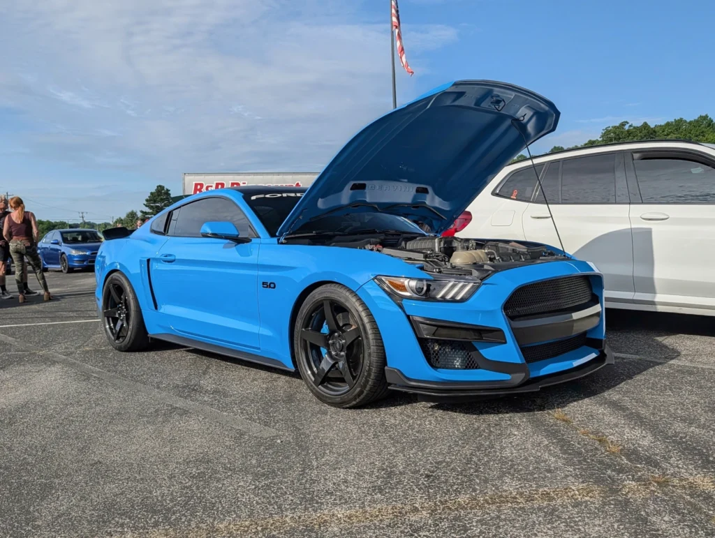 Blue Ford Mustang with hood open and visible engine, parked beside a white SUV in a car event setting