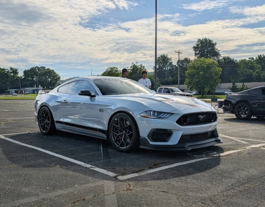 Silver Ford Mustang GT parked in an outdoor lot with two people standing behind it under a partly cloudy sky