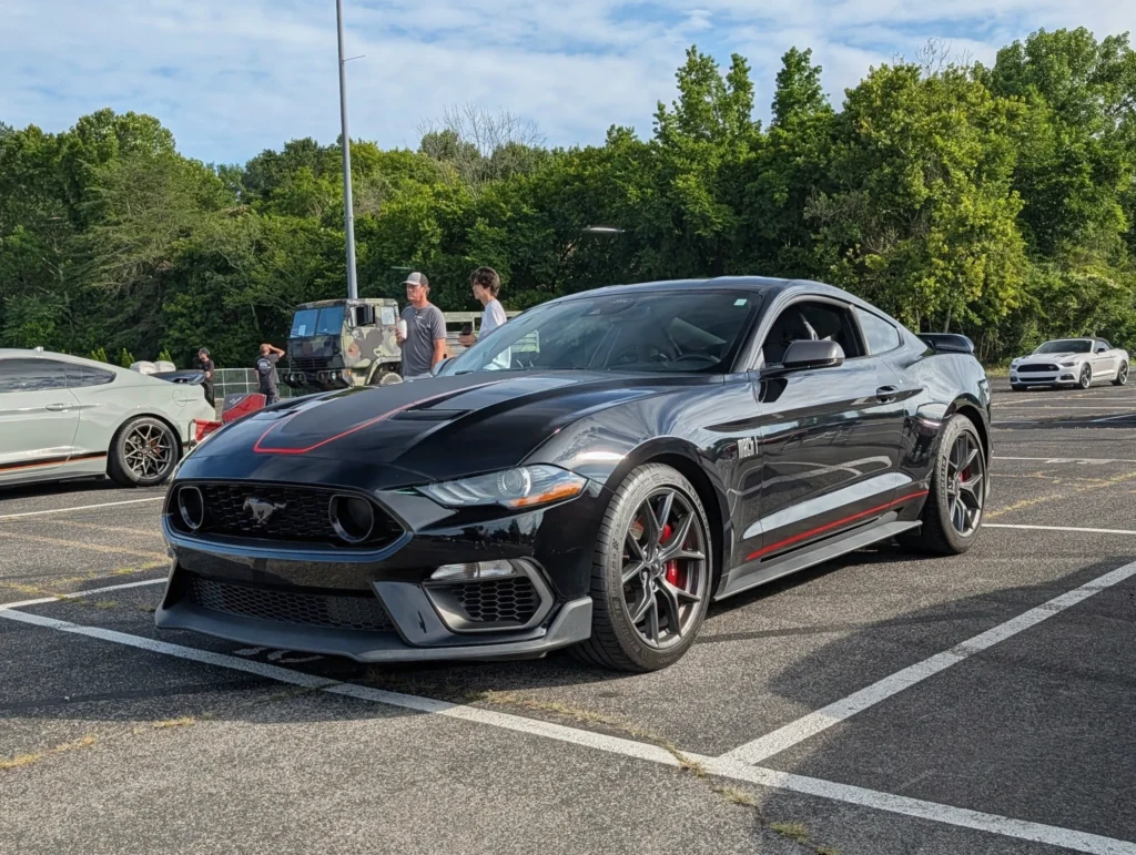 Black Ford Mustang Mach 1 with red accents parked at an outdoor car event with other vehicles and people nearby