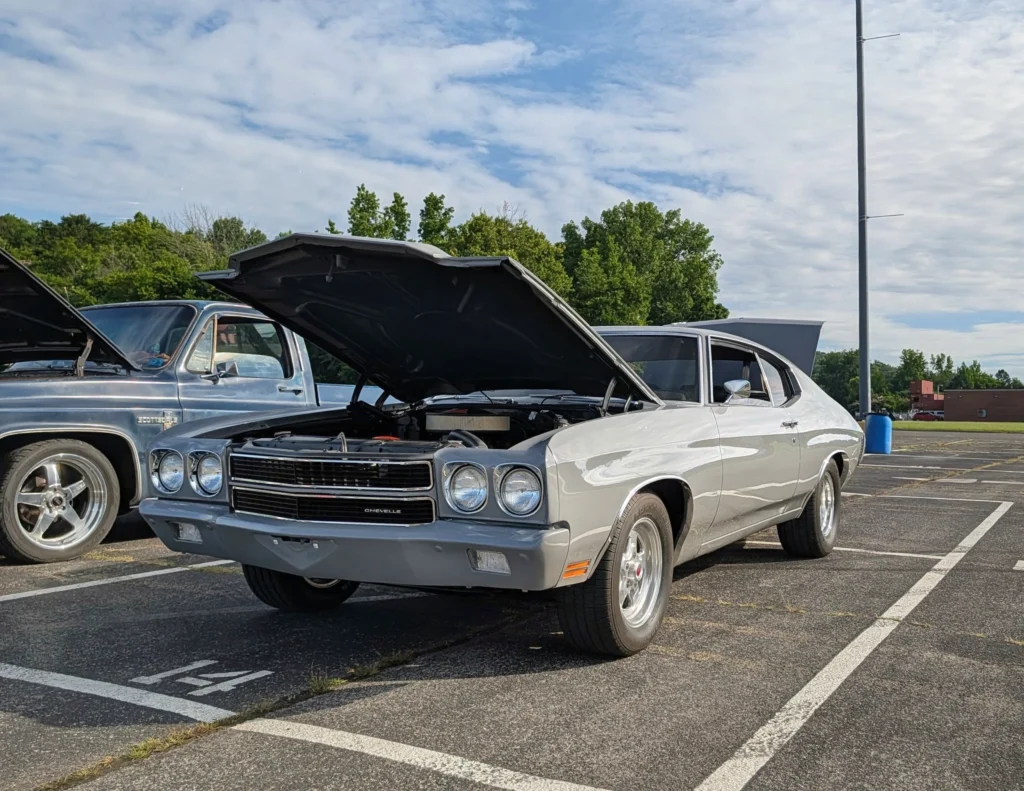 White Chevrolet Chevelle with hood open in an outdoor parking lot alongside a blue Chevrolet Scottsdale pickup truck.