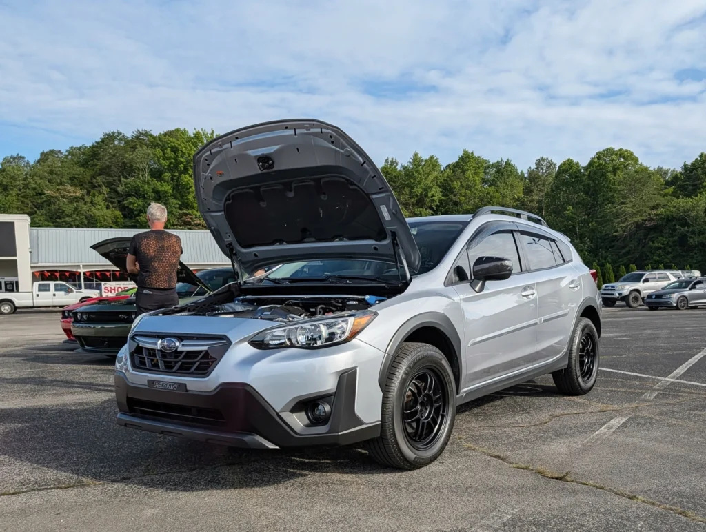 Silver Subaru Crosstrek with hood open in a parking lot at a car meet with trees and other cars in the background