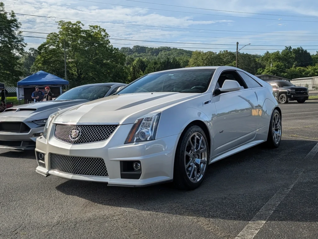 White Cadillac CTS-V coupe parked at an outdoor car event with other vehicles and tents in the background