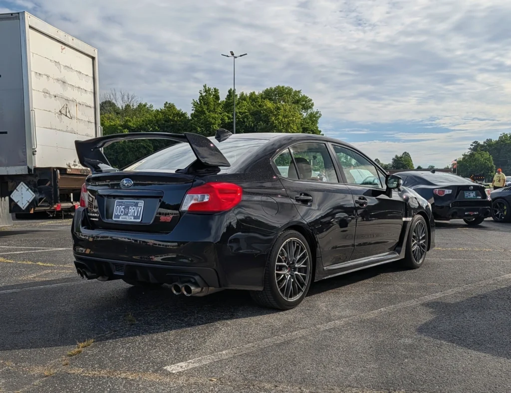 Black Subaru WRX STI sedan with rear wing parked in a lot with a white trailer and trees in background
