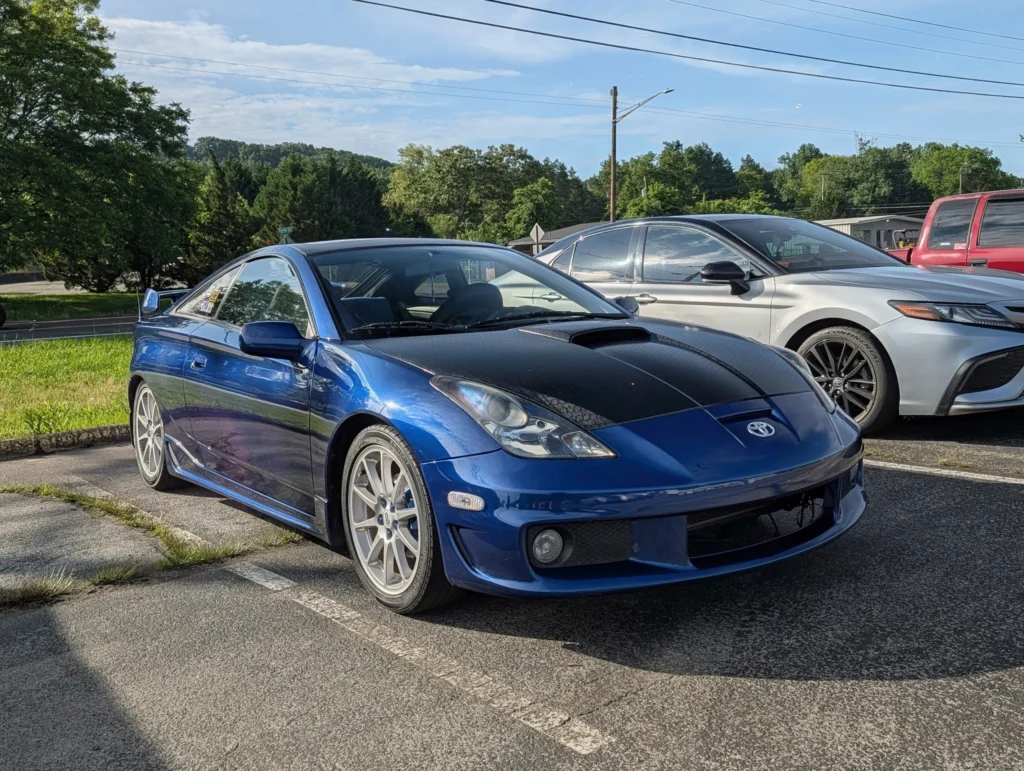 Blue Toyota Celica with black hood parked near a white Toyota sedan in an outdoor lot on a sunny day