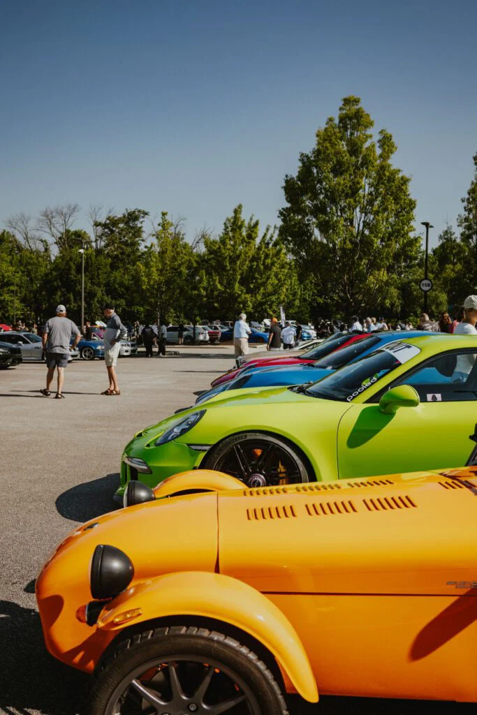 Row of colorful sports cars including an orange Caterham Seven and a lime green Porsche 911 parked at an outdoor car event with people wal