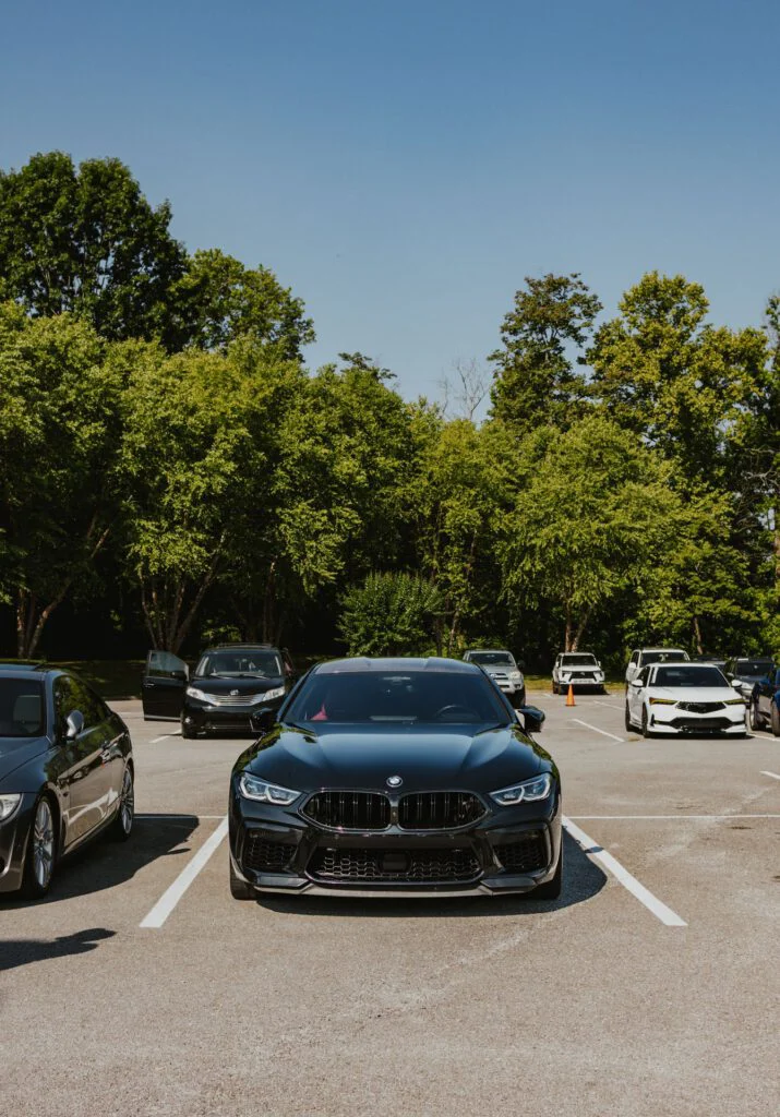 Black BMW M8 Competition Gran Coupe parked in a lot with other vehicles and trees in the background under a clear sky