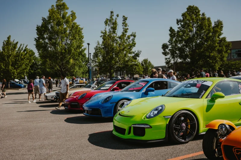 Row of colorful Porsche 911s parked outdoors with people walking around at a car event under clear skies.