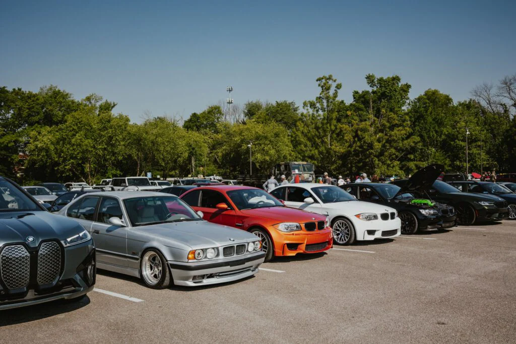 Line of parked BMW vehicles including silver E34, orange E82, white E82, and black E90 models in a car event parking lot.