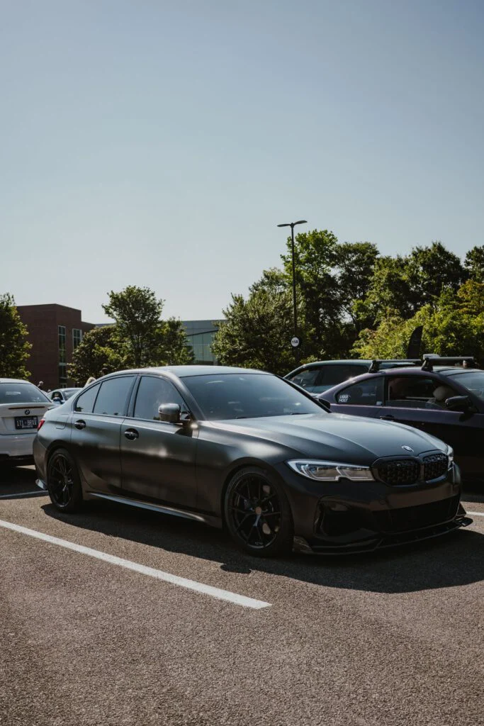 Matte black BMW M340i G20 parked in a lot with other vehicles and trees in the background under clear sky.