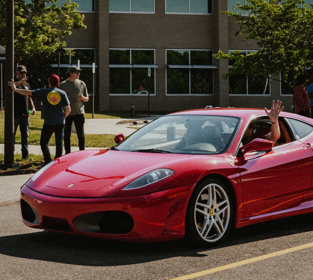 Rosso Corsa Ferrari F430 parked outdoors with three people in background and a hand waving from inside the vehicle