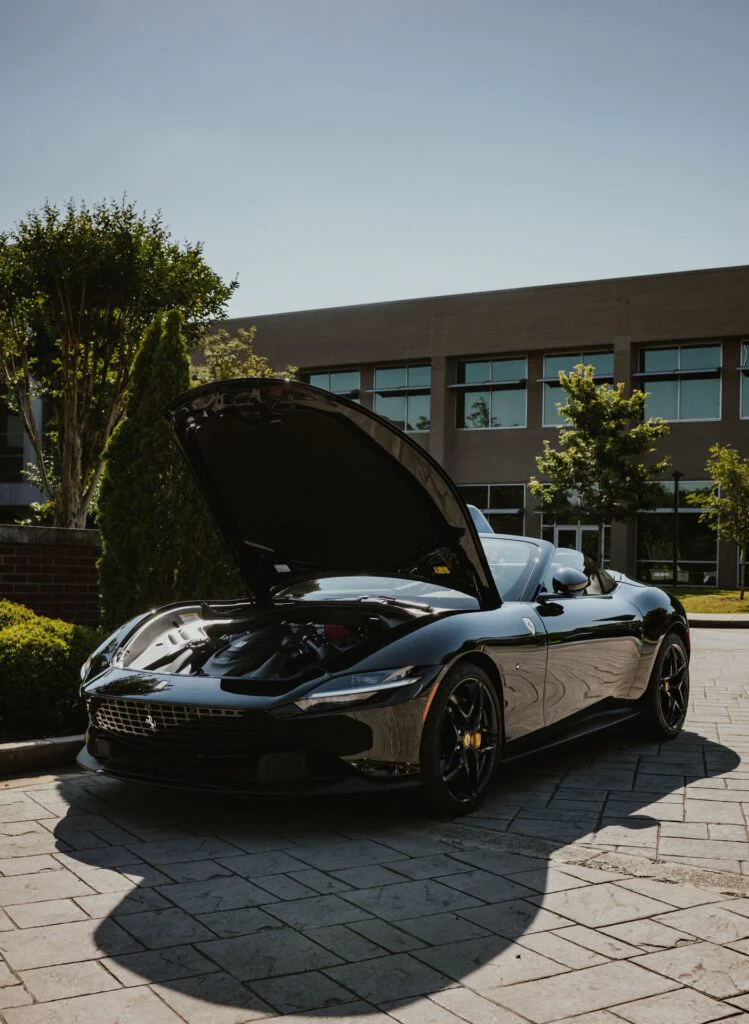 Black Ferrari Roma Spider with its front trunk open in an outdoor paved area near a building and trees