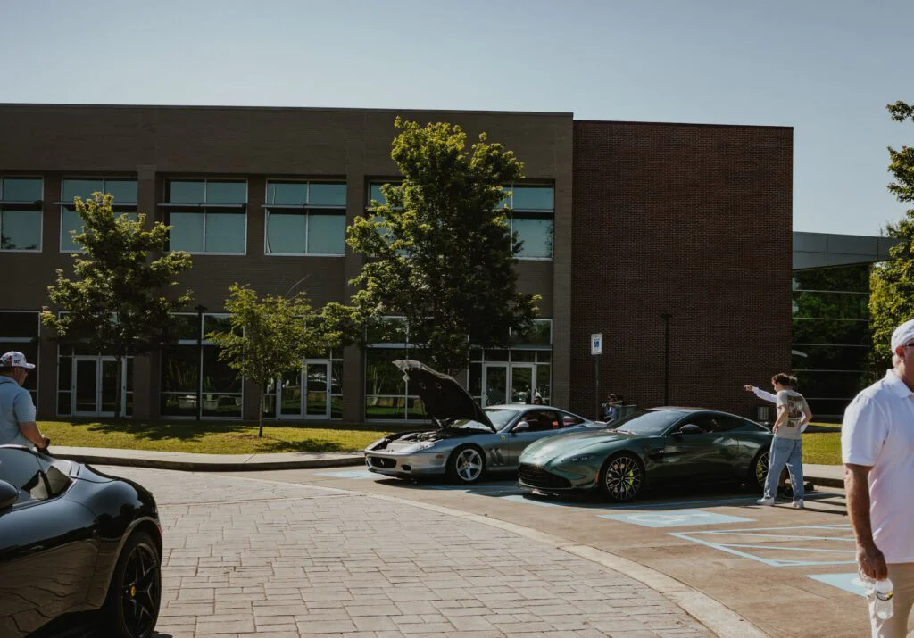 Silver Ferrari 550 Maranello with open hood and green Aston Martin Vantage F1 Edition parked outside a brick building during a car event