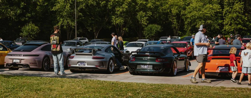 Group of Porsche 911s in various colors parked outdoors with people standing and walking nearby at a car event