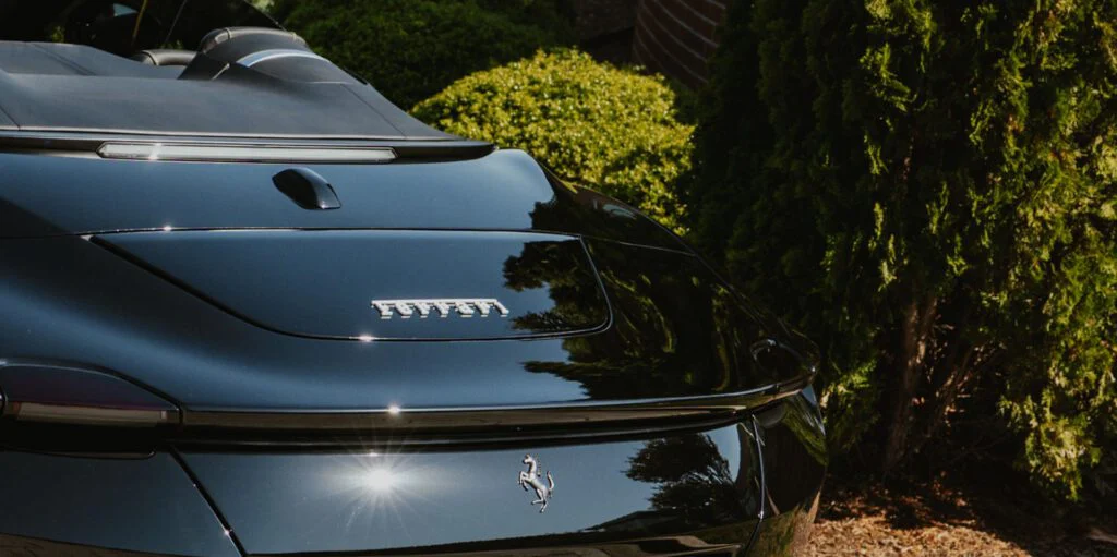 Rear view of black Ferrari Roma Spider with top down, parked near green bushes and a brick wall
