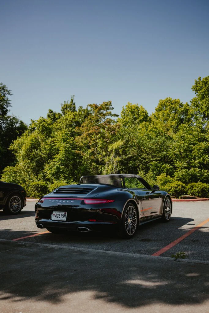 Black Porsche 911 convertible parked in a lot with green trees in the background under clear sky