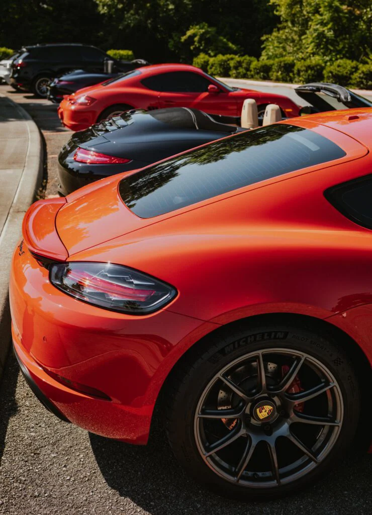 Rear section of an orange Porsche 718 Cayman in a parking lot with several other cars, including red and black Porsche models