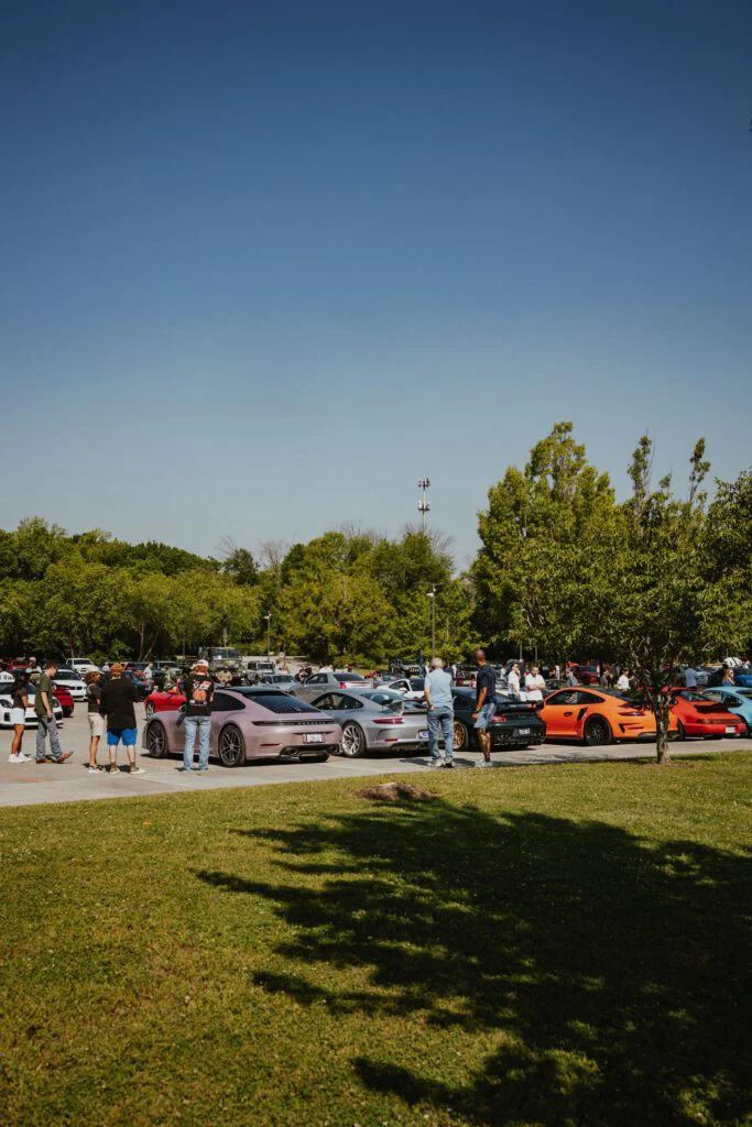 Group of people gathered around several Porsche 911s parked in an outdoor lot with trees in the background.
