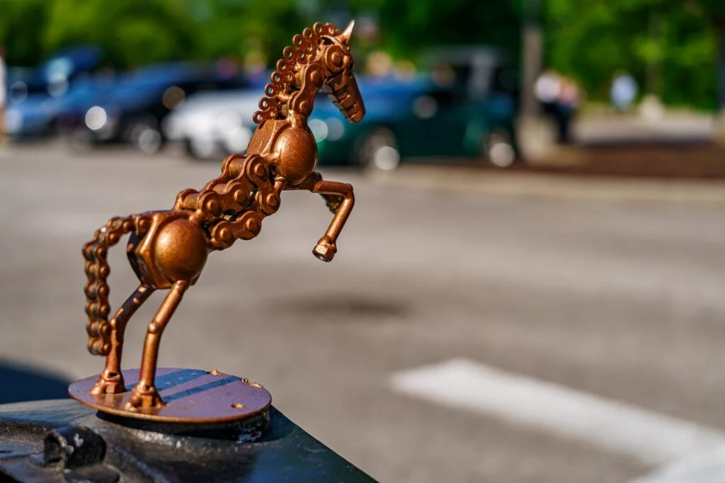 Rust-colored metal horse hood ornament with blurred parked cars in background