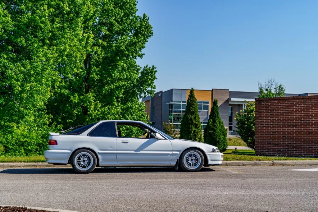 White Acura Integra parked on pavement with green trees and a modern building in the background