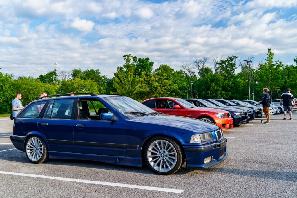 Blue BMW E36 Touring parked at a car meet with other cars and people in the background under a cloudy sky
