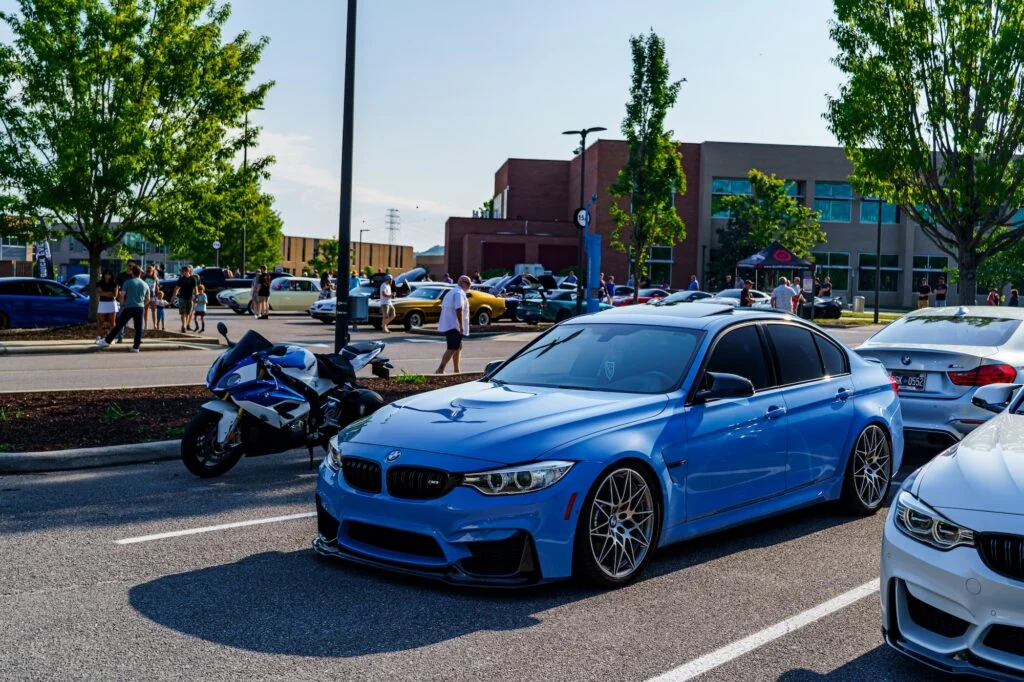 Blue BMW M3 and white BMW M4 parked in a lot with other cars and people at a car meet on a sunny day