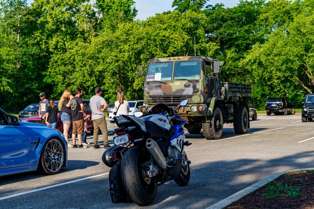 Camouflage military truck parked in a lot with a group of people nearby and a blue motorcycle in the foreground
