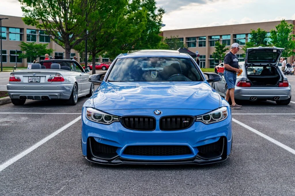 Blue BMW M3 parked in a lot with silver BMW convertible and silver hatchback, man standing with hatchback trunk open