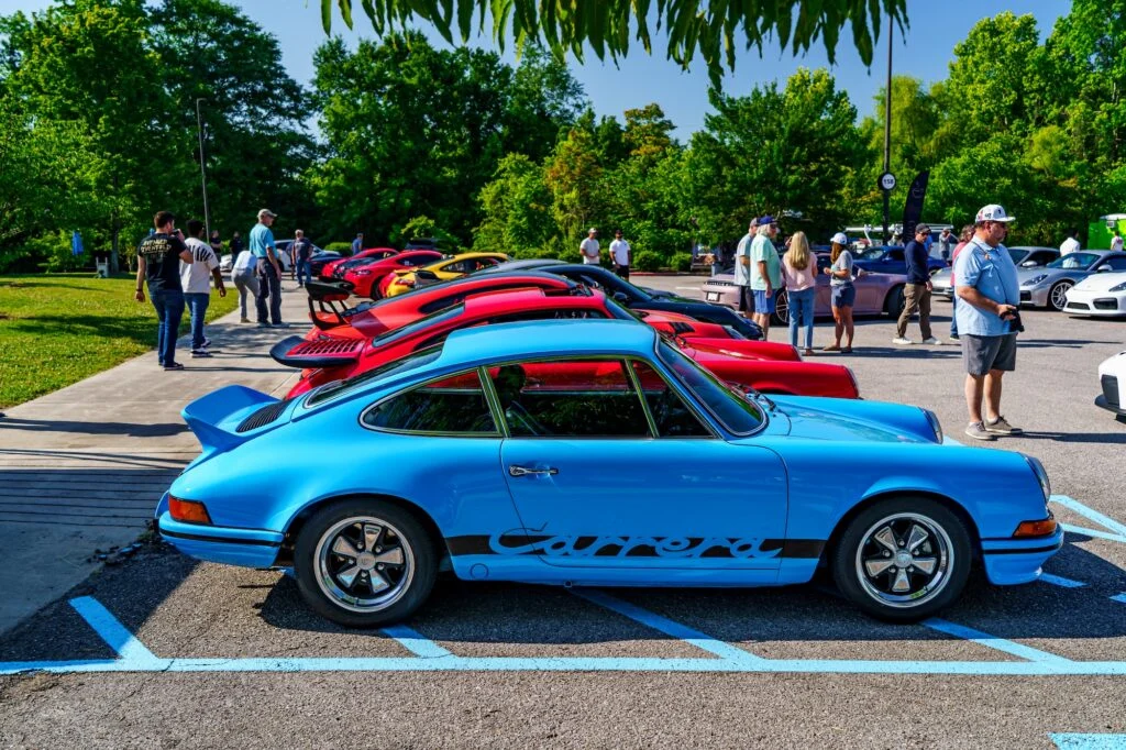 Row of parked sports cars including a blue Porsche Carrera, people walking and standing around in a parking lot at an outdoor