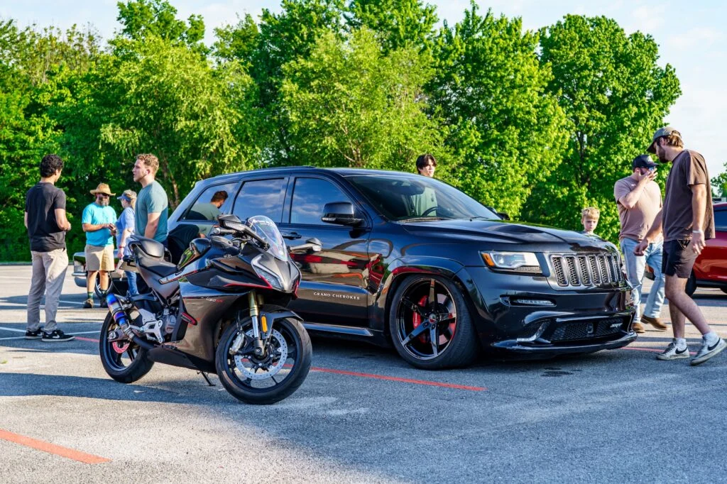Black Jeep Grand Cherokee SRT and black CFMOTO 450SS parked outdoors with several people standing nearby under trees