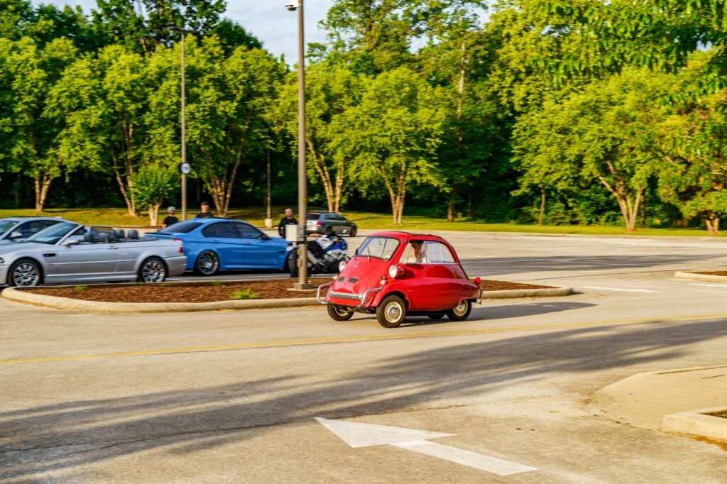 Red BMW Isetta microcar driving on a street near parked sports cars and motorcycles with trees in the background