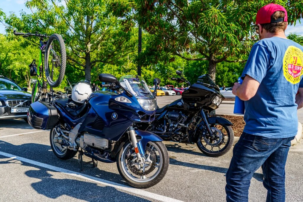 Blue BMW K1200RS with a bicycle mounted on back and black motorcycle parked nearby in a sunlit lot with trees and a man ne
