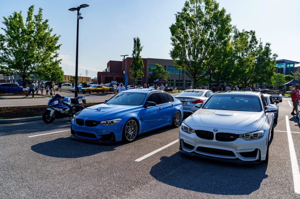 Blue and white BMW M3 and M4 sedans parked side by side in a parking lot at a car meet with people and trees in the background